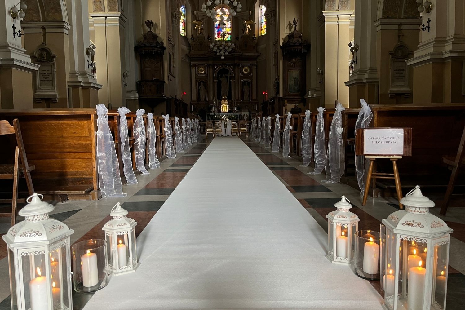 Biały dywan w kościele udekorowanym na ślub, z lampionami i tiulem przy ławkach