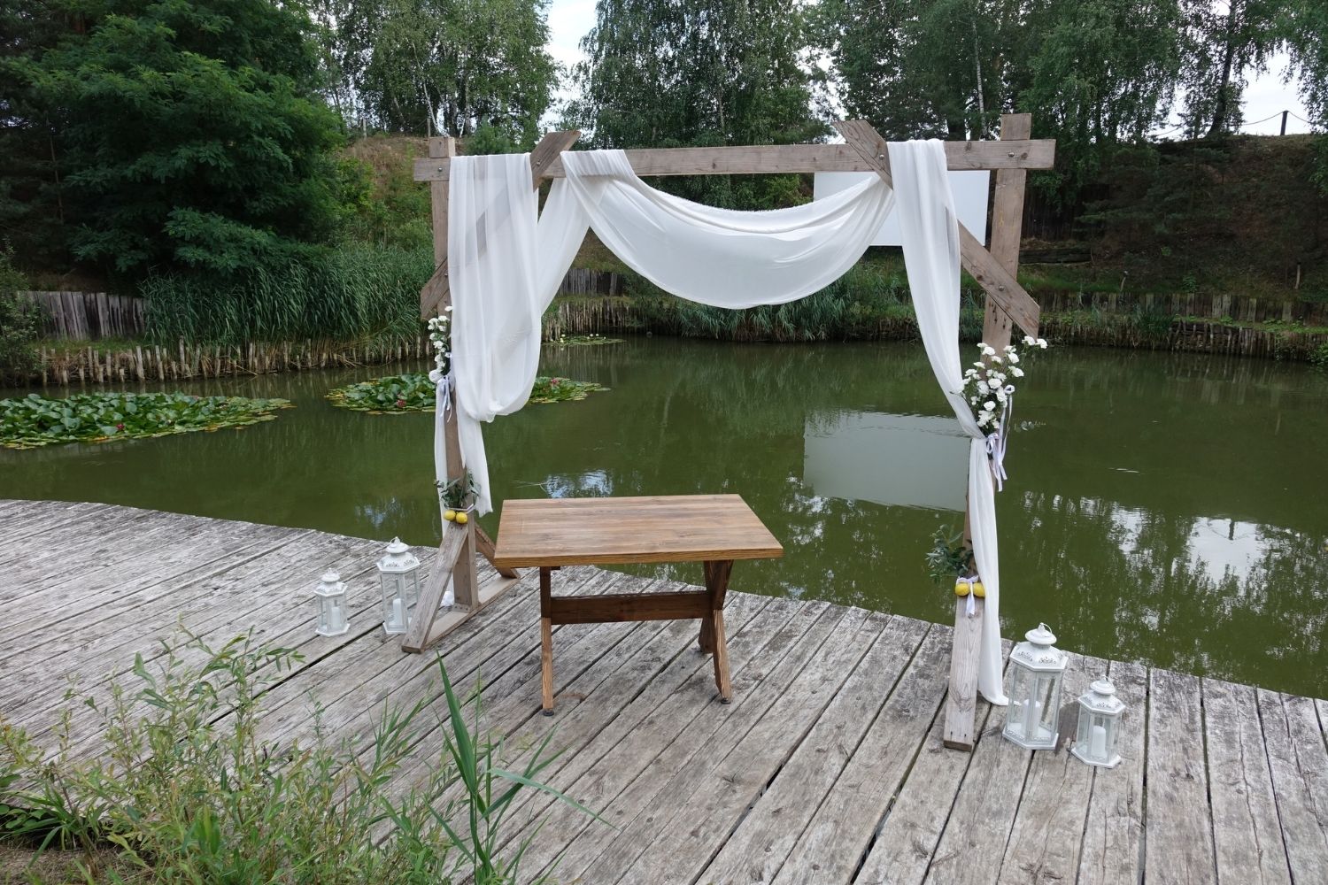 Dekoracja ślubna plenerowa, drewniany łuk z białymi zasłonami i kwiatami nad jeziorem