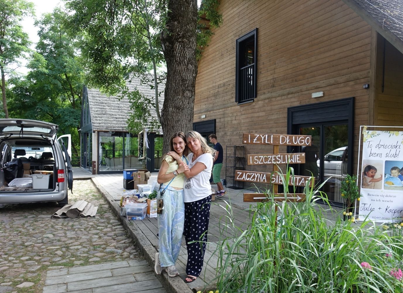 Duet Majowe Chwile - Maja i Kasia podczas montażu dekoracji ślubnych przed drewnianą salą weselną.