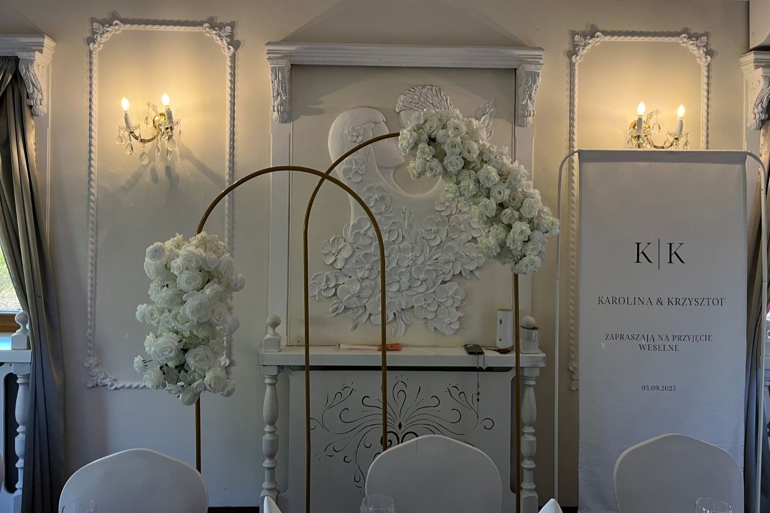 Elegancka dekoracja ślubna, dwa złote łuki z białymi różami i tablica powitalna dla pary młodej.