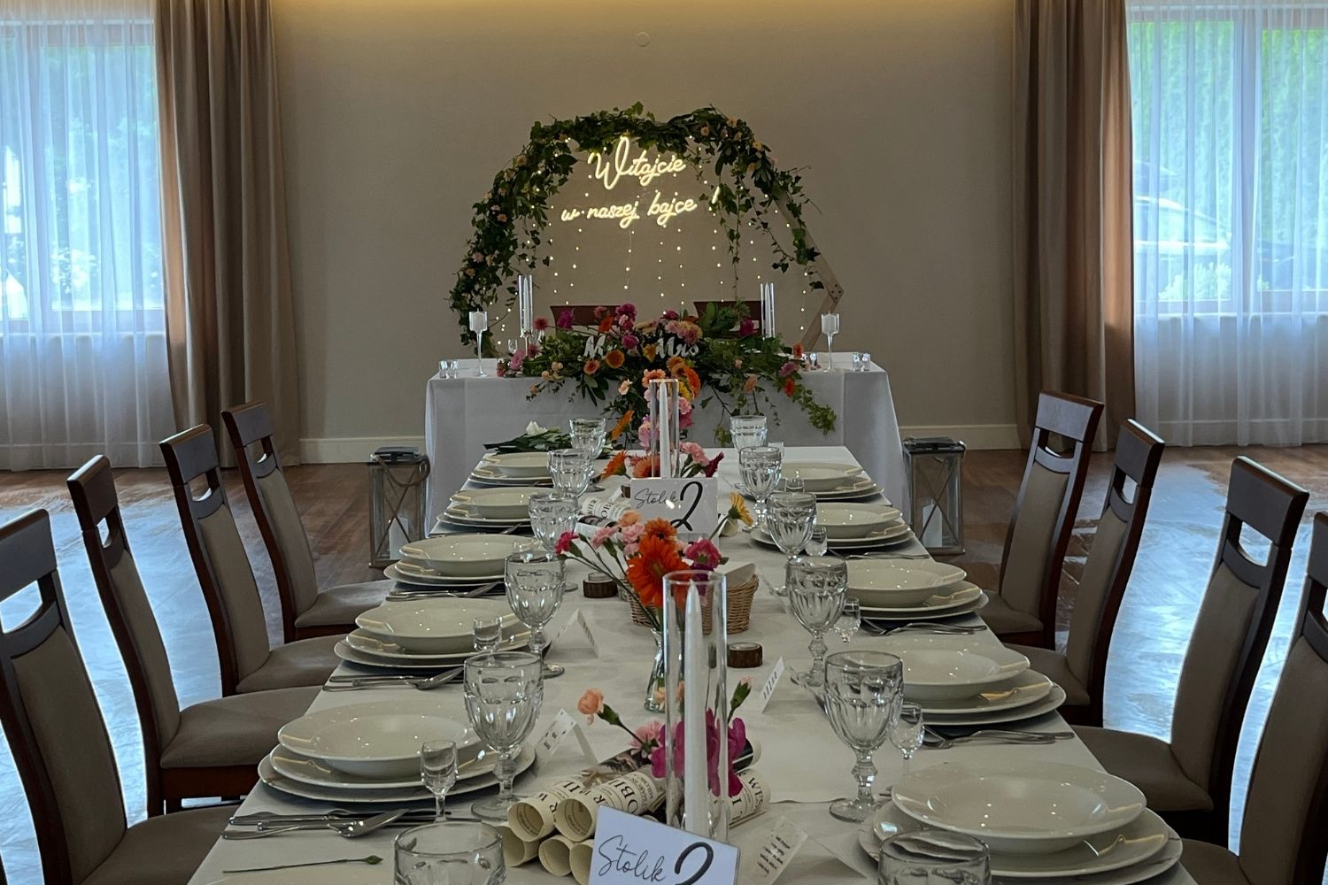 Dekoracja stołu gości na weselu, w tle stół pary młodej ze ścianką w kształcie heksagonu i neonem "Witajcie w naszej bajce".