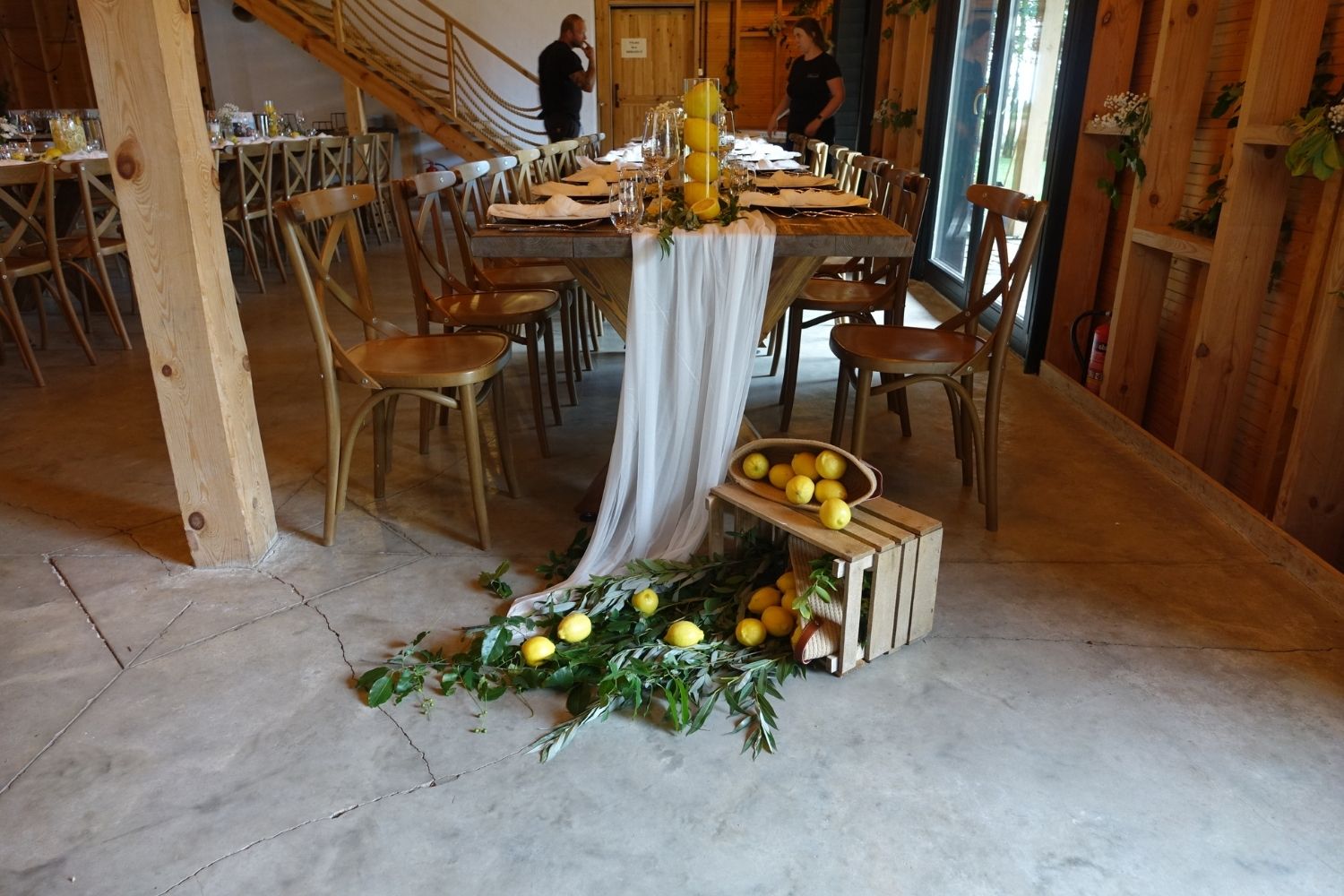 Rustykalna dekoracja weselna, drewniana skrzynka z cytrynami i zielenią przy długim, drewnianym stole.